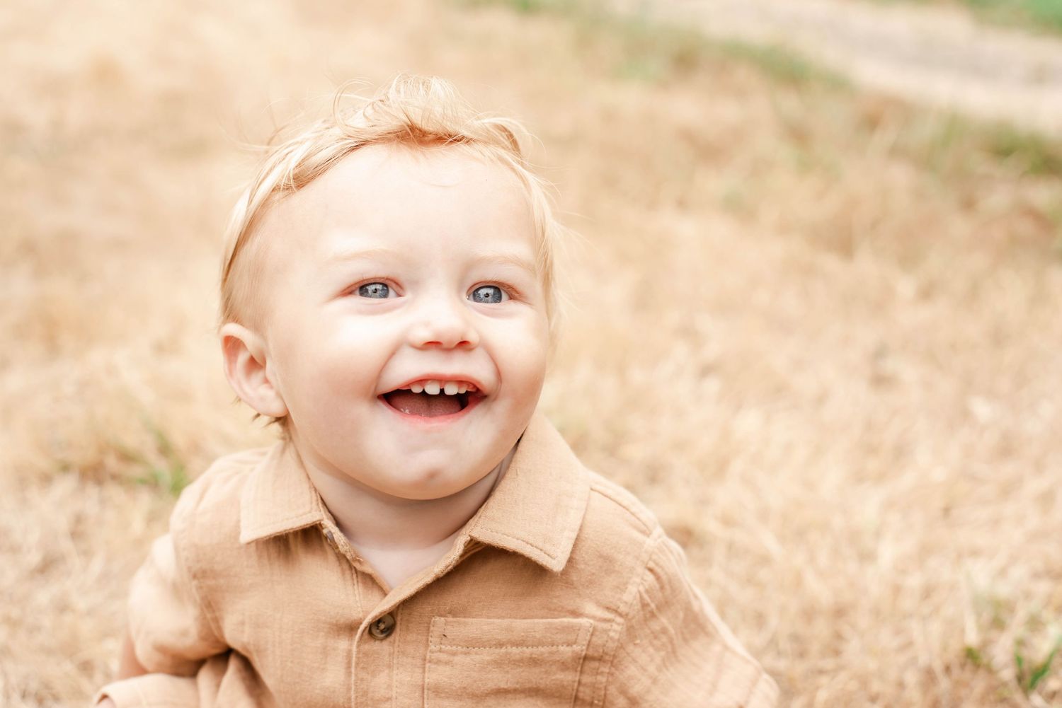 A smiling baby in a tan shirt sits outdoors in a grassy field on a sunny day.