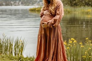 A pregnant woman in a flowing rust-colored maxi dress stands by a serene lake surrounded by yellow wildflowers.