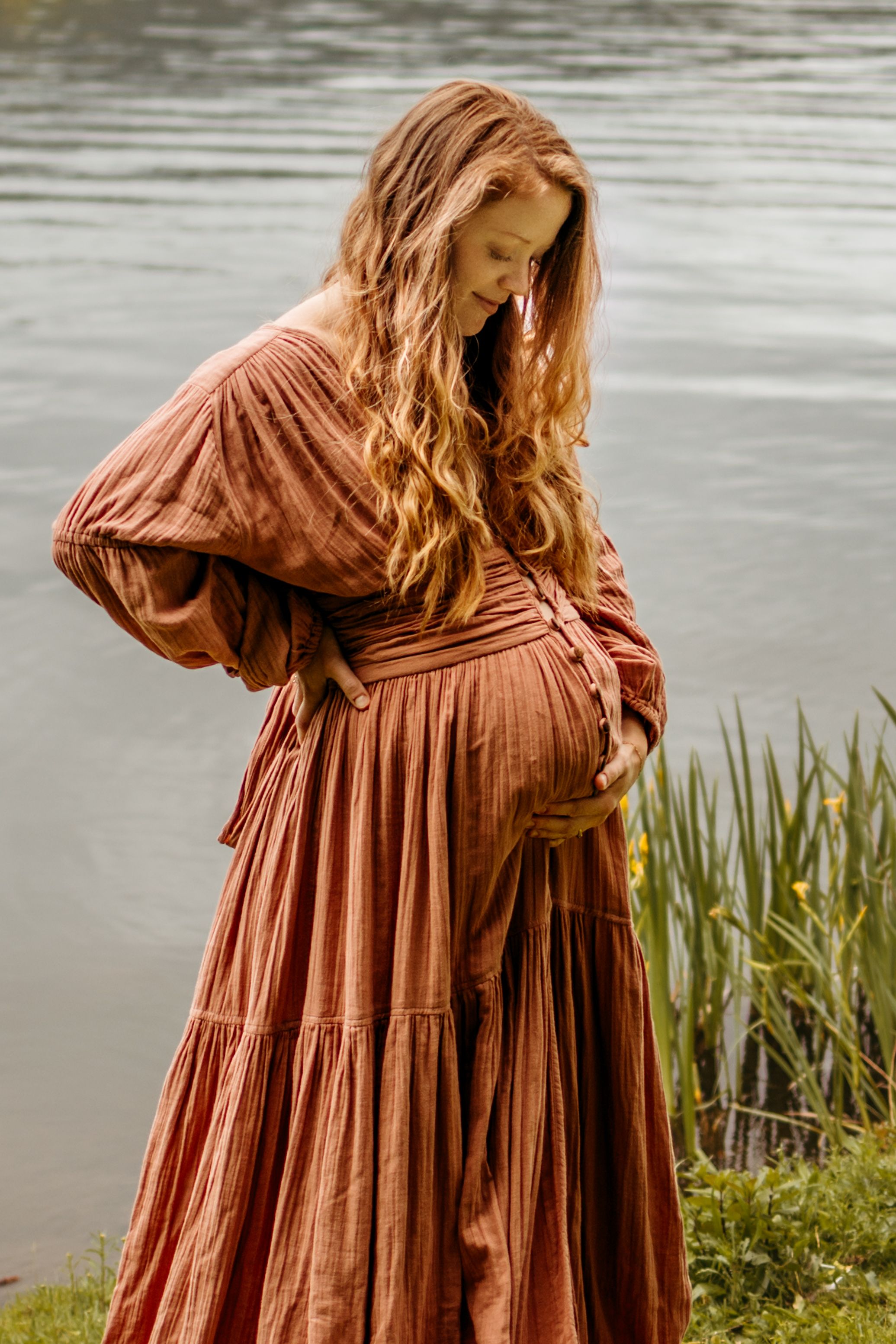 Person in flowing rust-colored tiered dress stands near water's edge with tall grass and reflective lake surface.