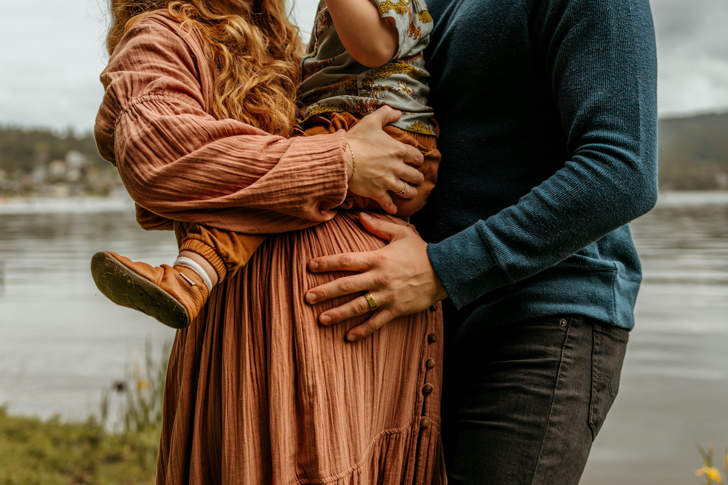 A couple embraces outdoors wearing a rust-colored dress and navy sweater against a moody lakeside backdrop.