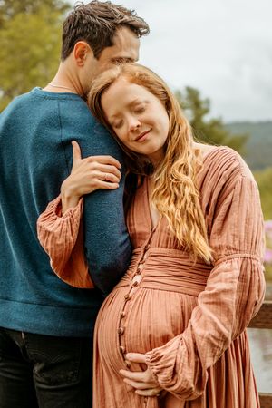 A couple shares an intimate embrace outdoors, with the person in peach-colored dress cradling their pregnant belly.