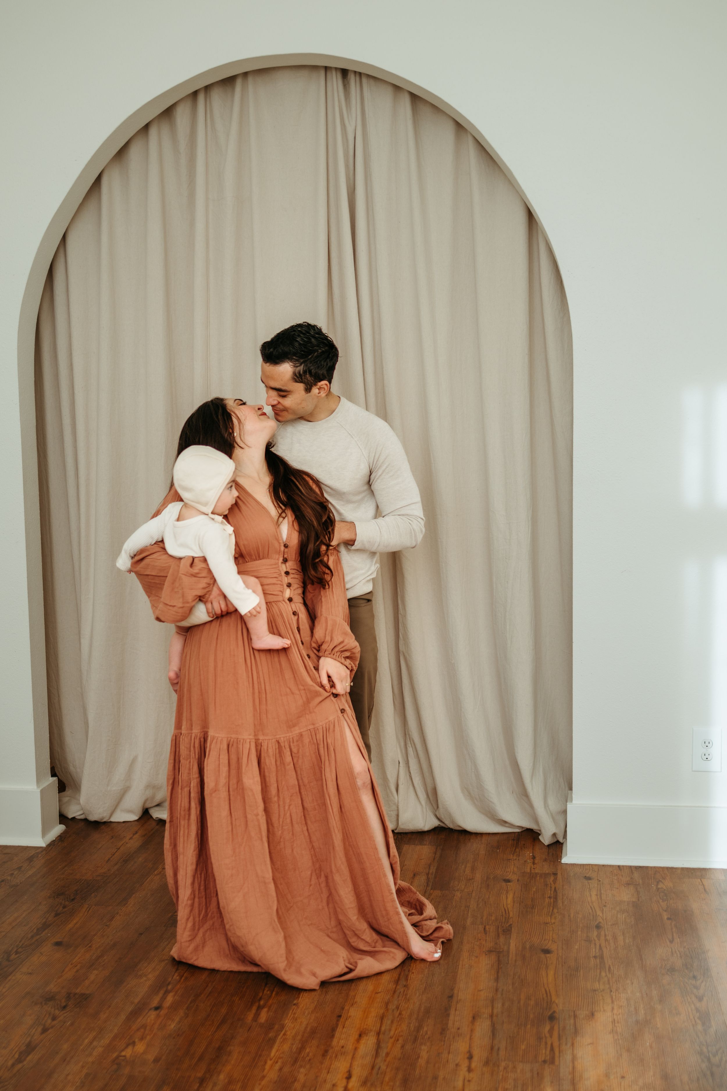 A couple in white and rust-colored clothing share playful poses against a beige curtain backdrop in a photo series.