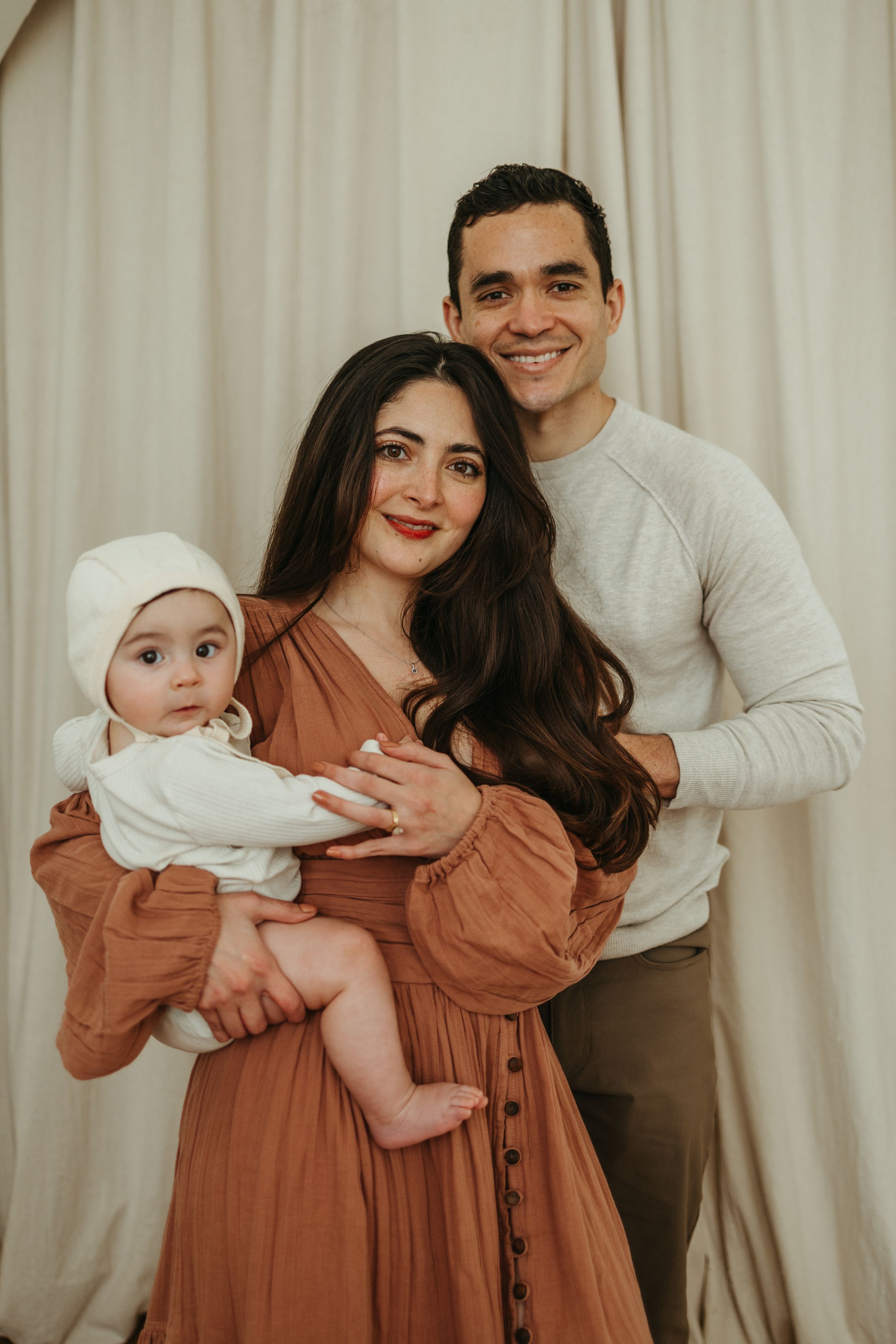A family portrait against a cream backdrop shows a couple in warm-toned clothing holding their baby dressed in white.