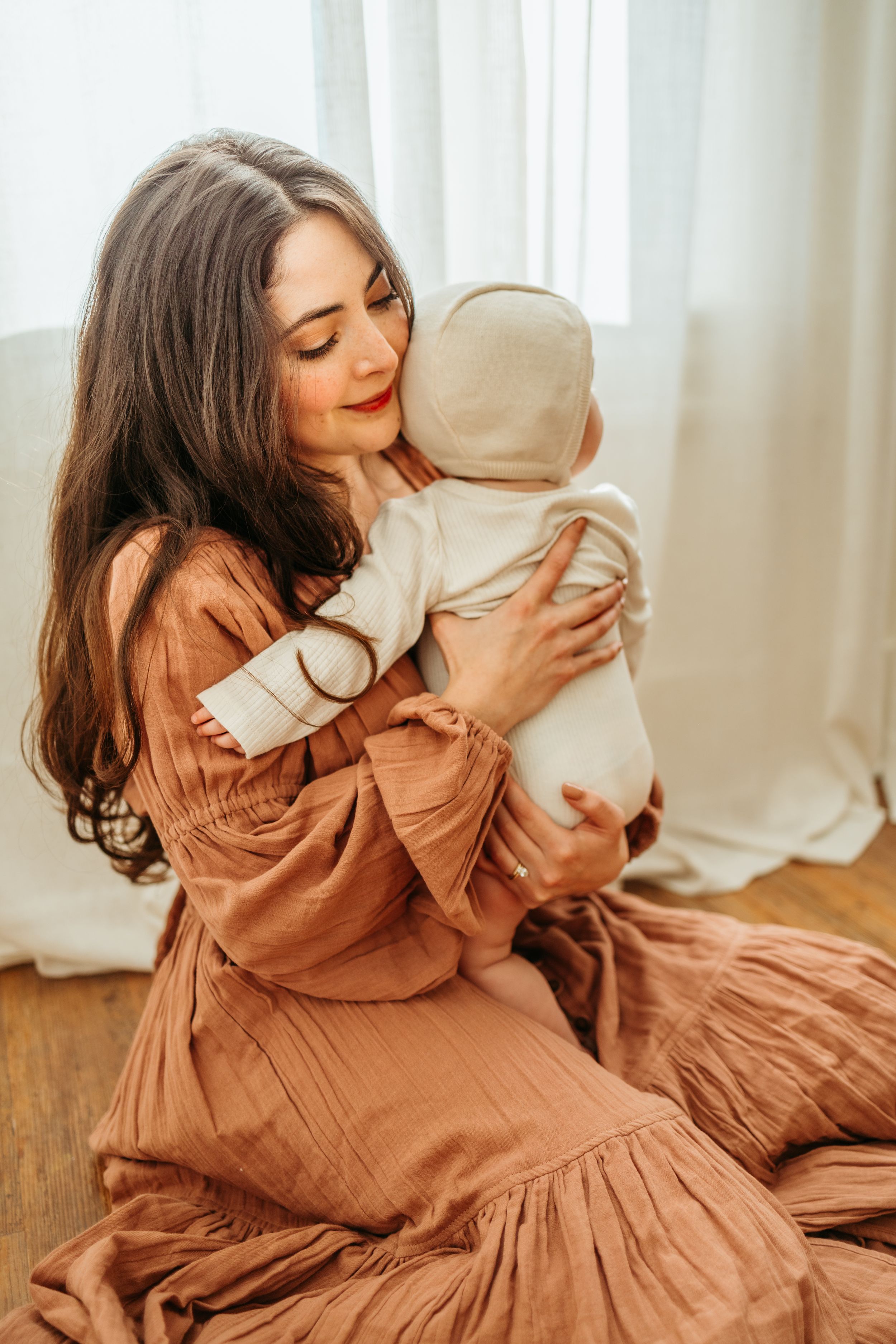 Mother in flowing rust-colored dress holds baby in white outfit by window in tender moment.