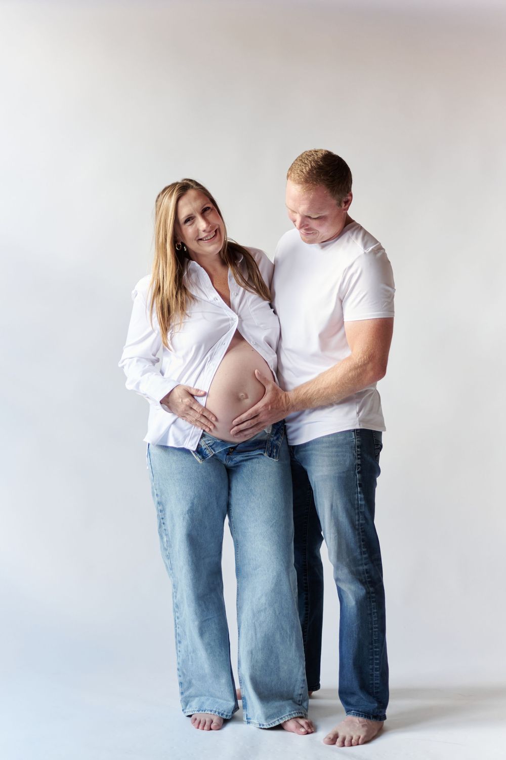 Expecting couple in matching white tops and jeans poses together for maternity photos against light background.
