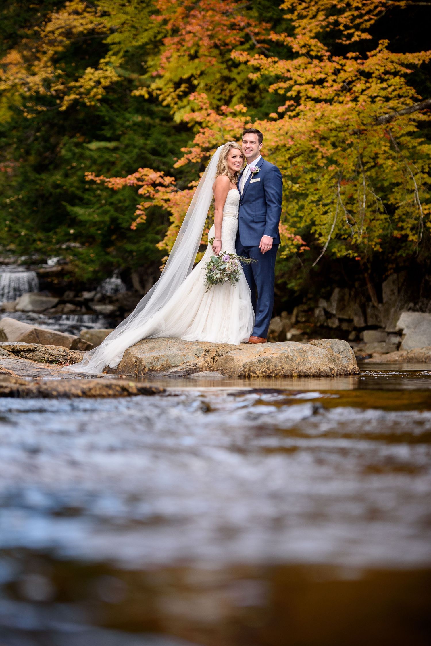 Couple in formal attire standing on rocks by a river with vibrant autumn foliage in the background.