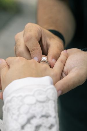 Close-up of hands placing engagement ring on finger with white lace sleeve.