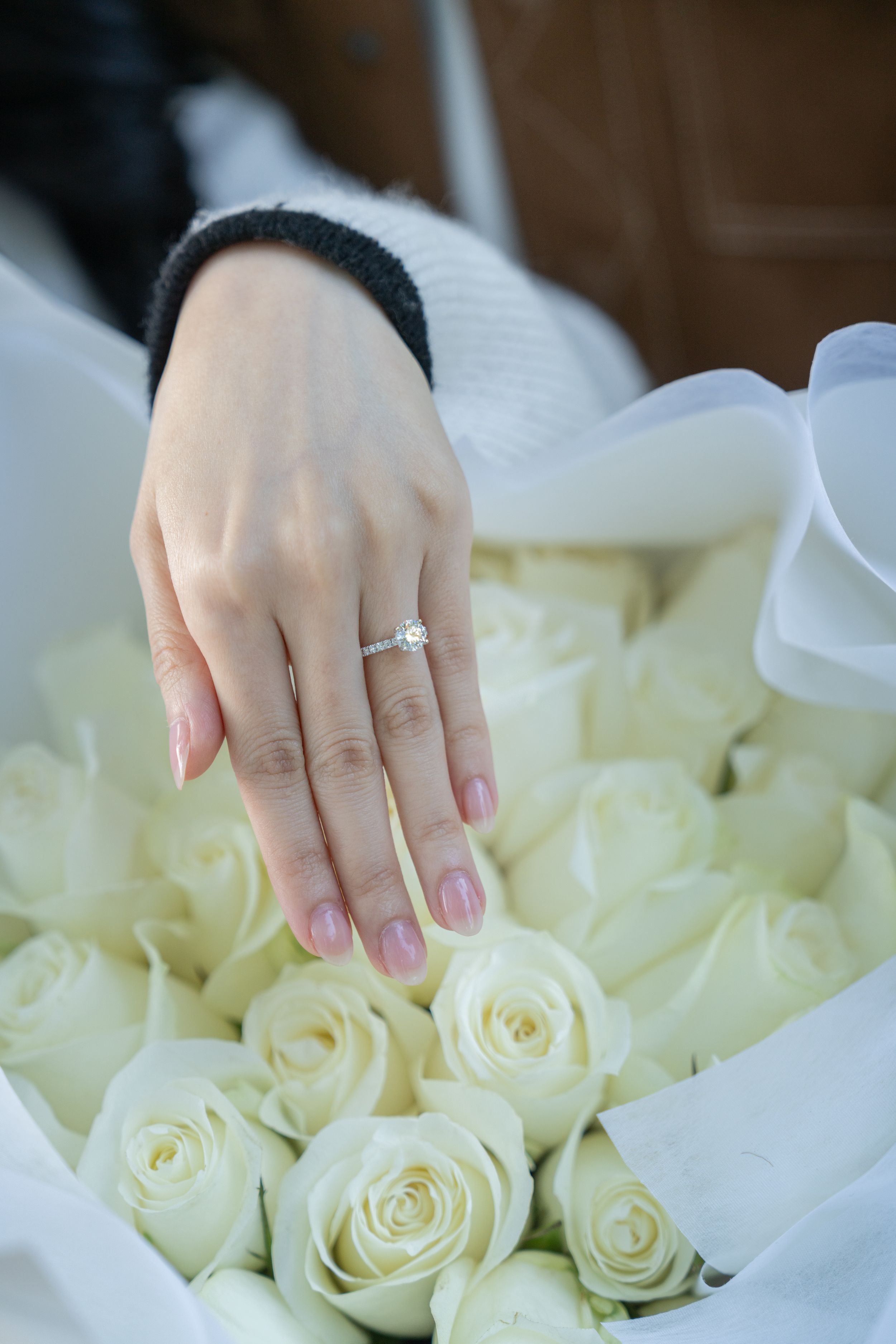 A close-up shot of hands displaying an engagement ring among white roses.