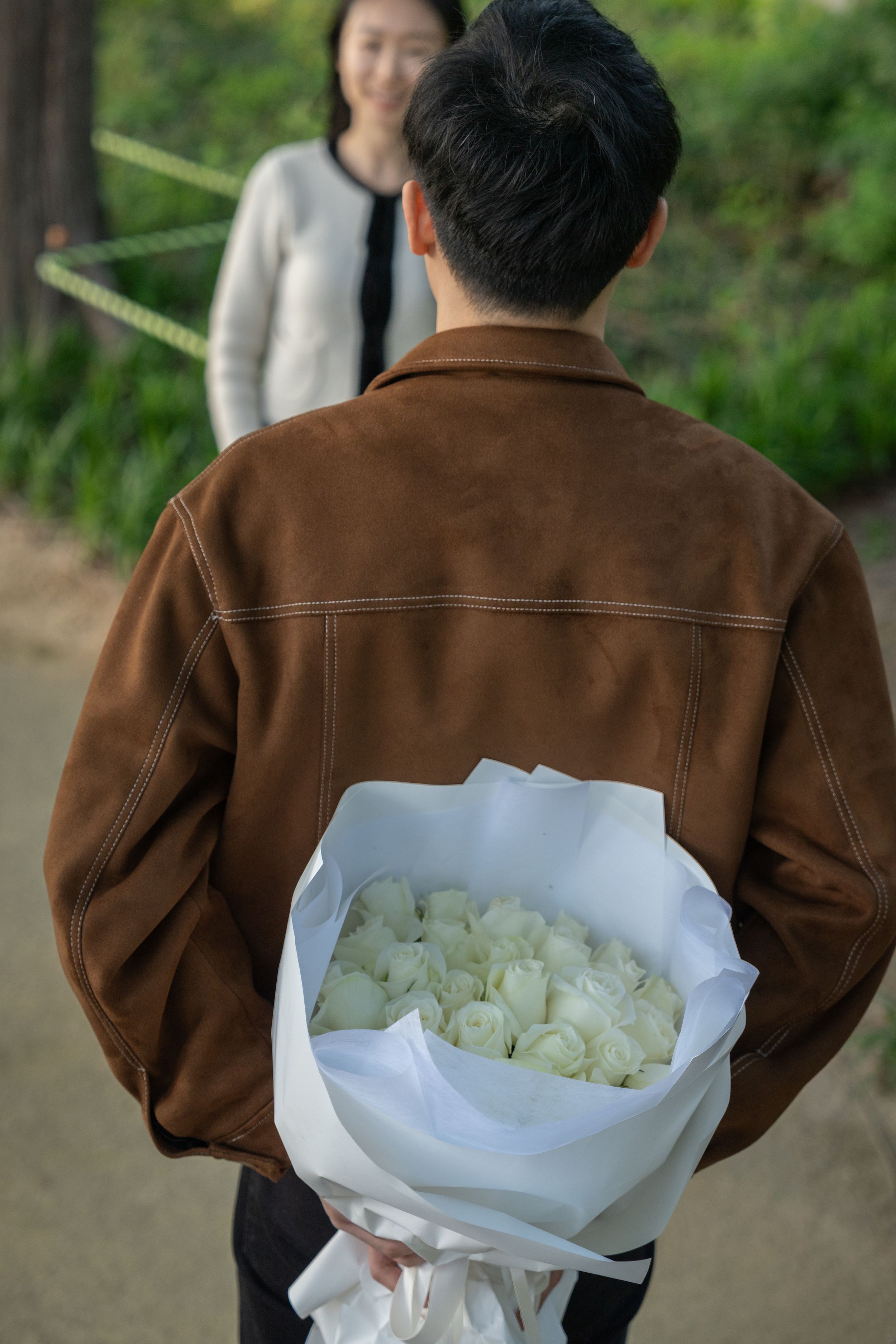 A person in a brown jacket holds a bouquet of white roses wrapped in paper.