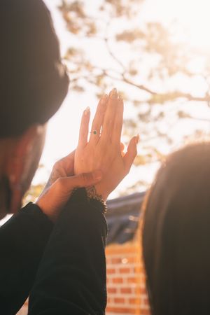A silhouetted person raises their hand against a bright sunset sky with lens flare and brick wall in background.