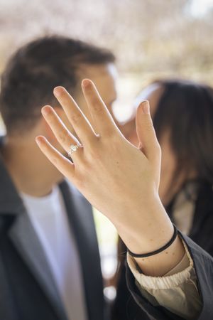 A hand displaying an engagement ring is raised against a blurred background.