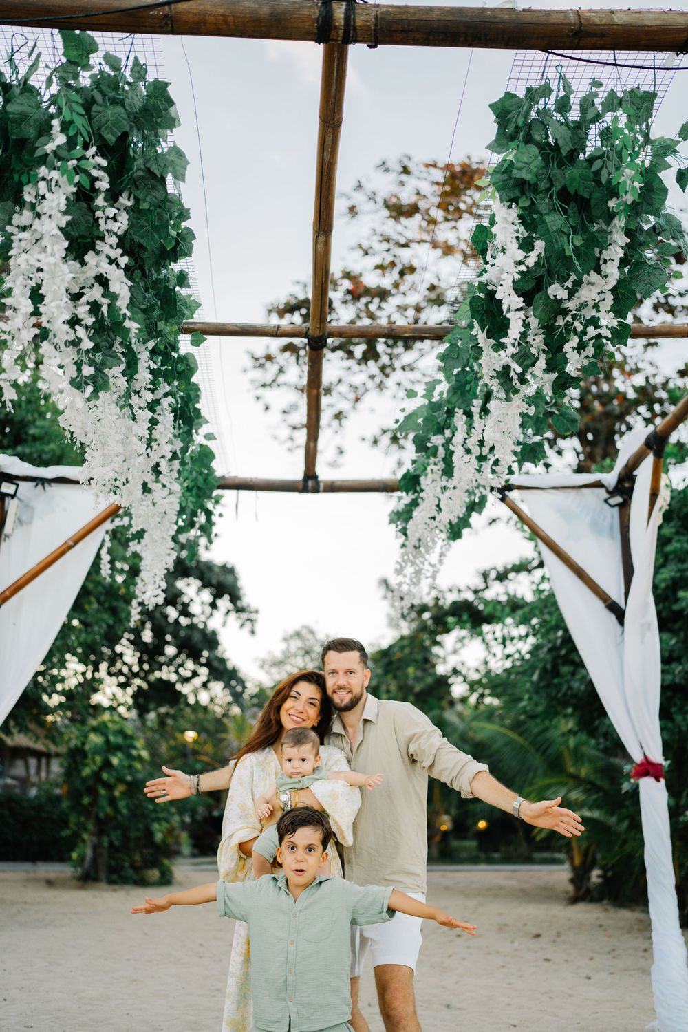 A family poses playfully under a decorated wedding arch with hanging greenery and white drapes.