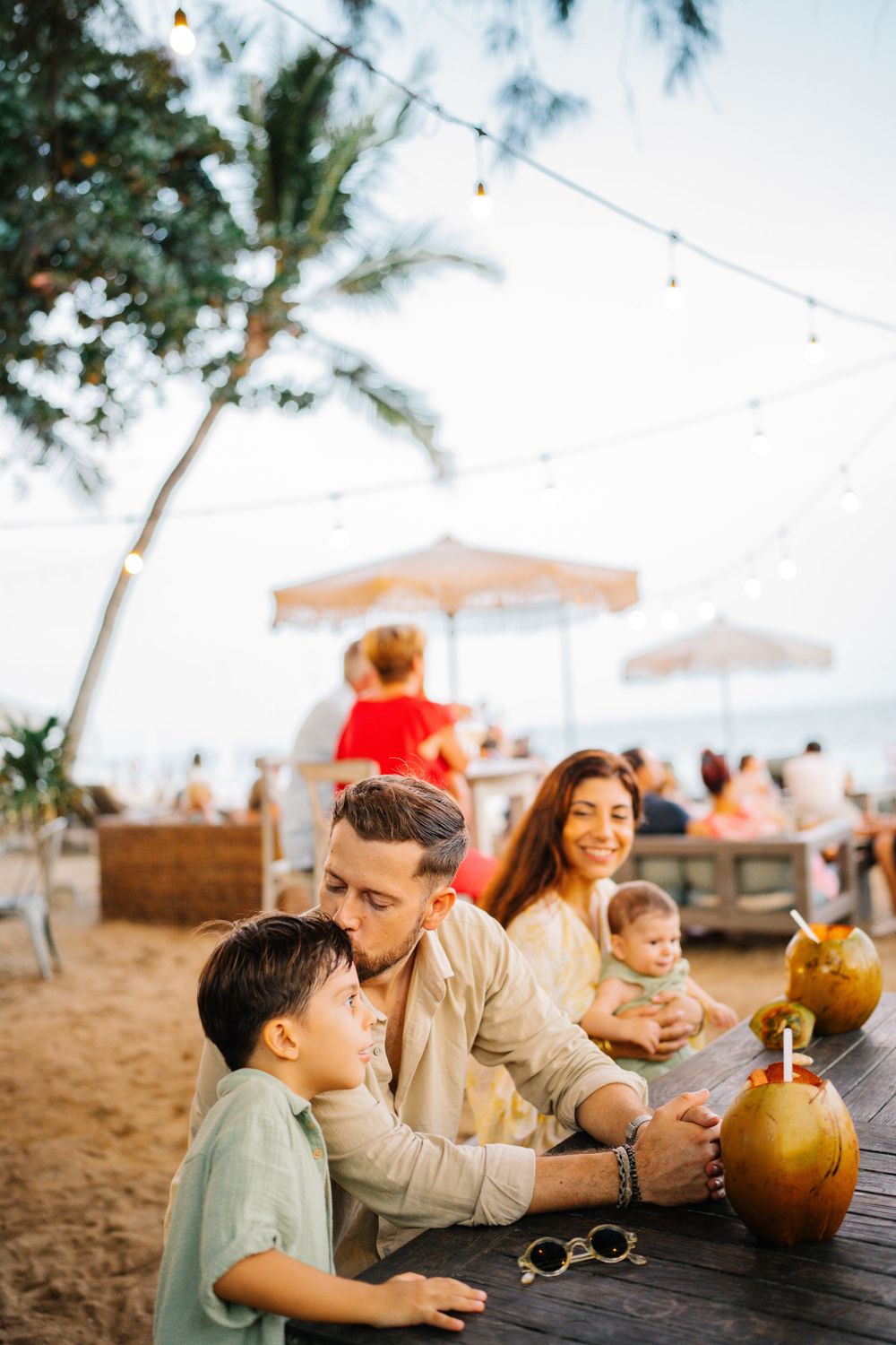 A family enjoys drinks at an outdoor beach restaurant with string lights overhead.