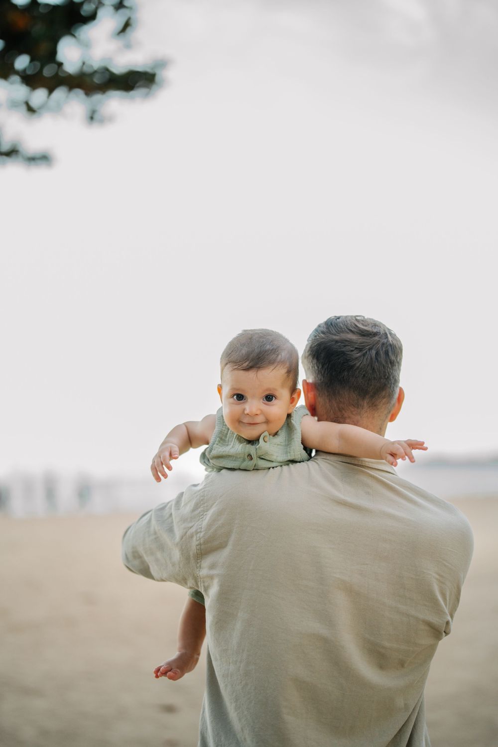 A parent holds a small child against a bright background while looking out at the ocean.