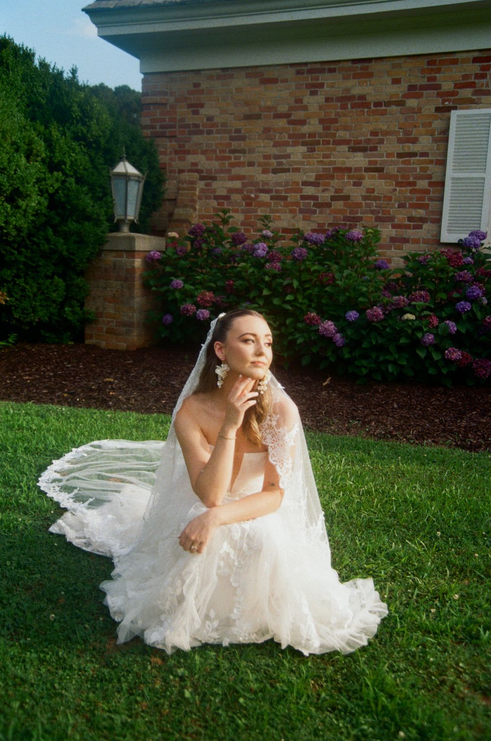 White lace wedding dress flows across green lawn with pink flowering bushes and brick house in background.