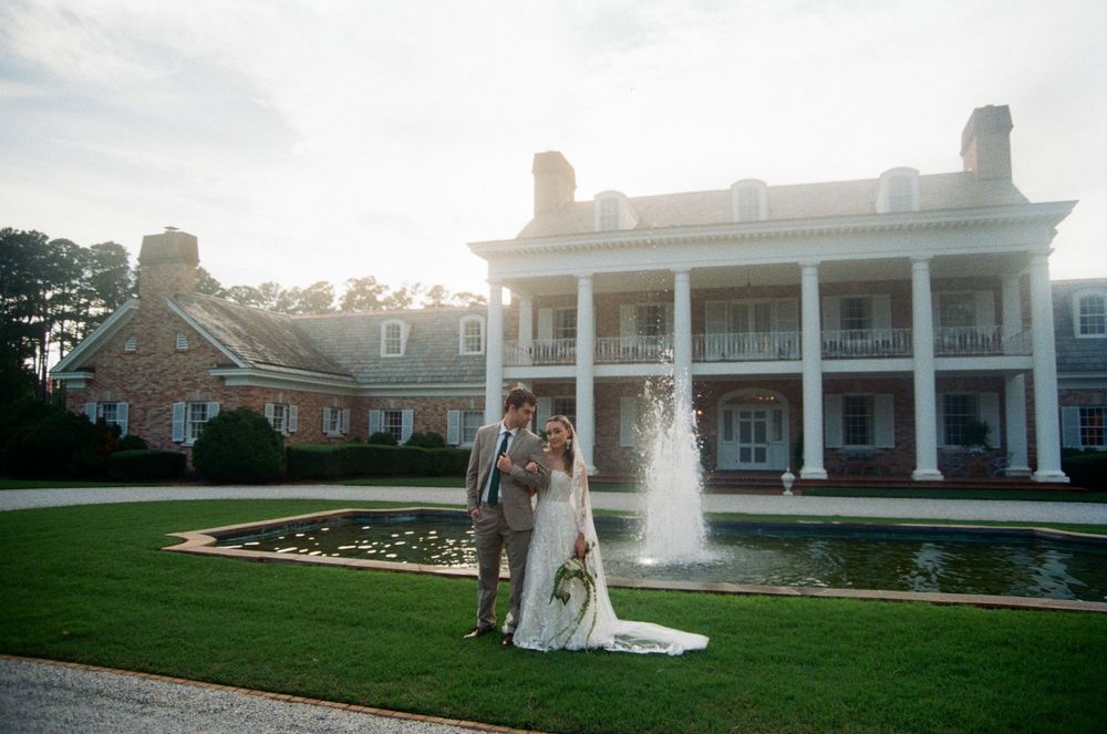 A wedding couple poses in front of a grand colonial-style mansion with white columns and a fountain on a manicured lawn.