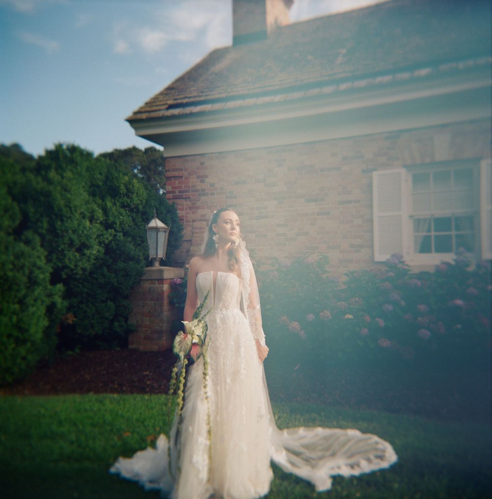 Long flowing wedding veil catches sunlight against brick building and green lawn.