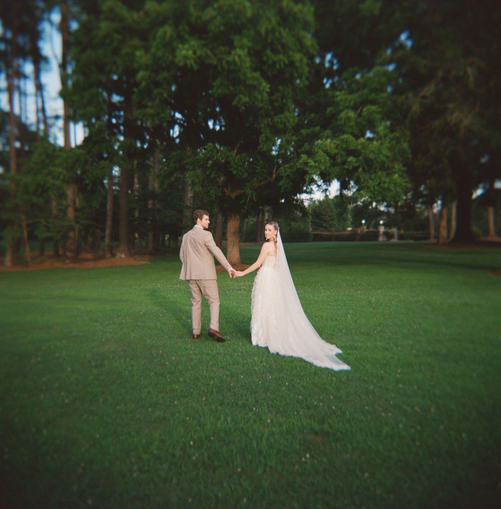 Couple in wedding attire walking across a scenic green lawn with tall pine trees in the background during sunset.