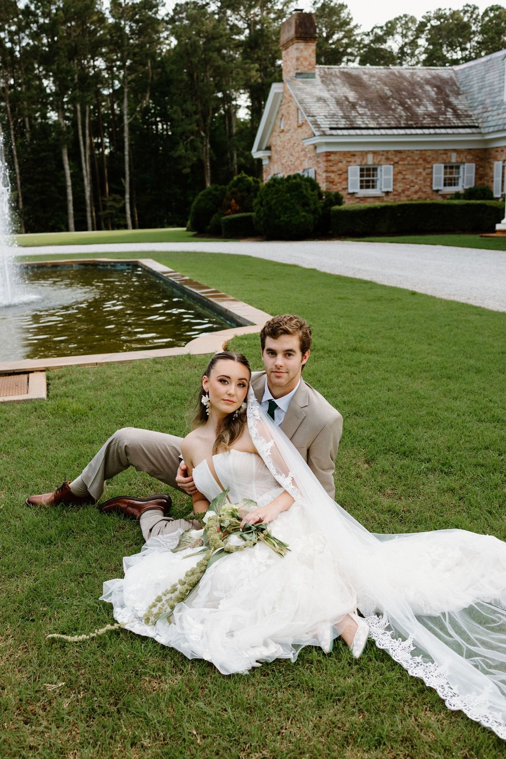 A couple in wedding attire laying intimately on grass near a rustic stone building on their special day.