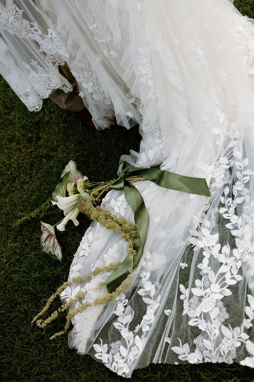 A delicate white wedding dress with lace details and floral appliques lies on grass with a small bouquet and green ribbon.