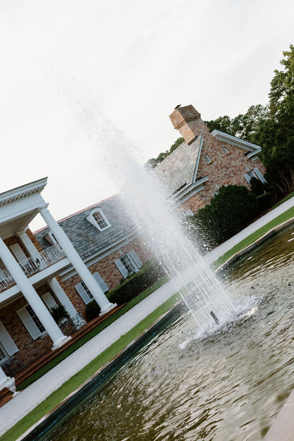A tall water fountain sprays in front of a colonial-style brick mansion with white pillars and a pond.