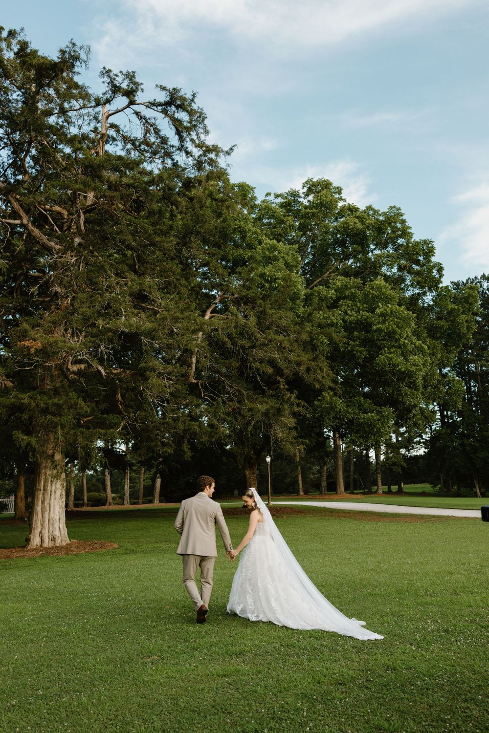 A couple in wedding attire walks together across a green lawn with tall oak trees in the background.