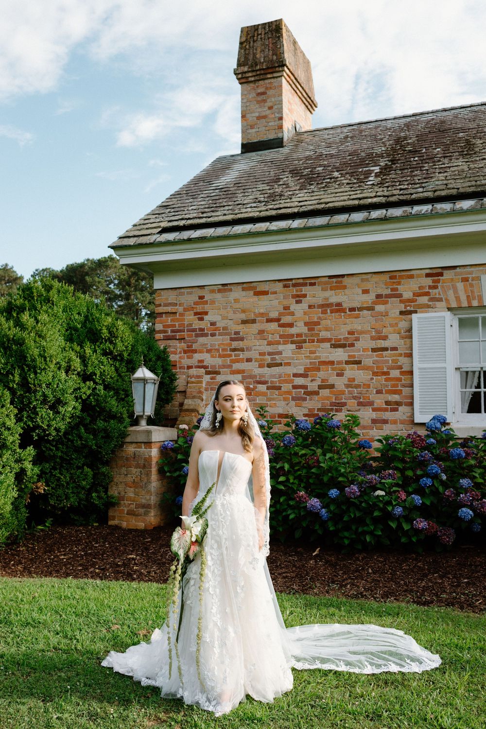 A beautiful wedding scene with billowing white dress against brick building with blue hydrangeas and manicured lawn.