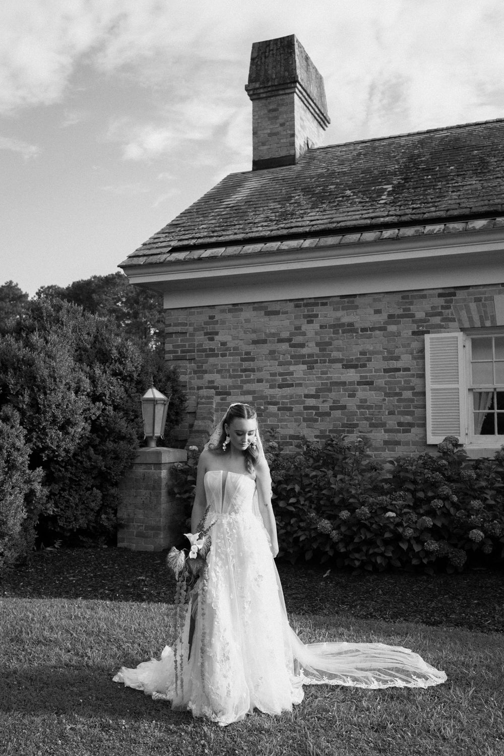 Black and white photograph of an elegant bride in a lace wedding gown with long train standing beside a brick building.