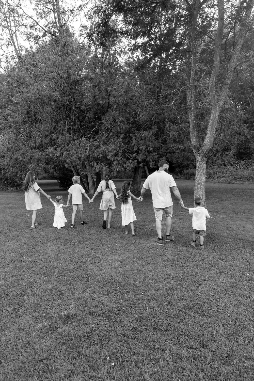 A black and white photo of a family of 8 walking away from the camera through a park on a beautiful late summer evening