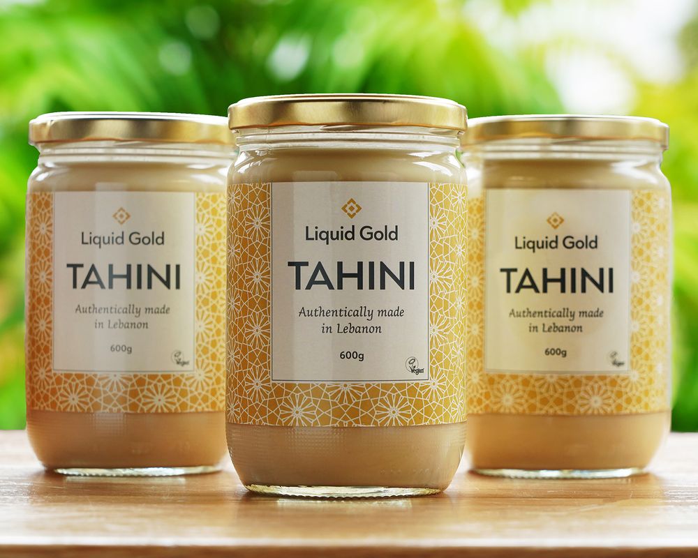 Three jars of Liquid Gold brand tahini displayed on wooden surface with green background.