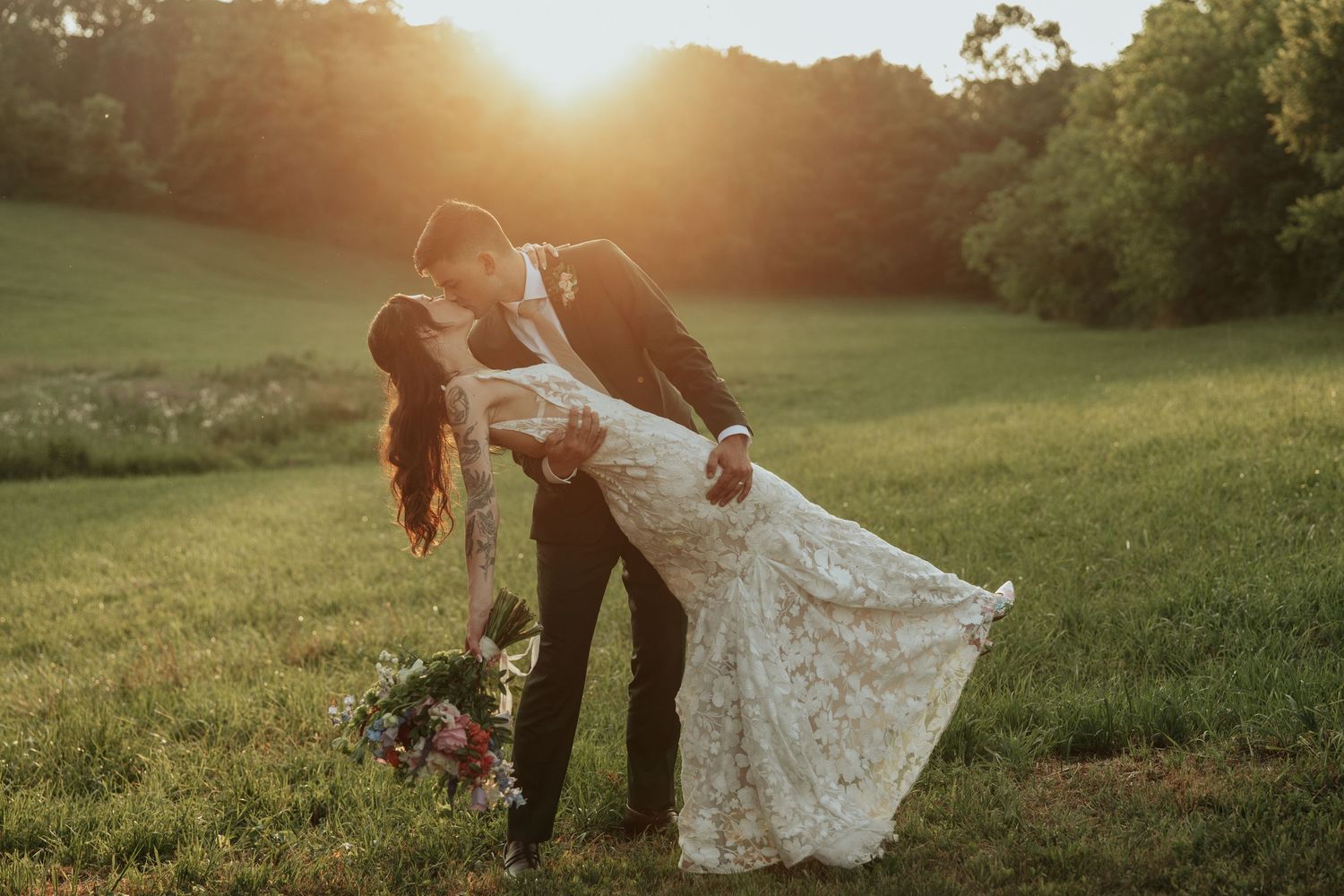 Golden hour sunset illuminates a grassy field as a couple celebrates their special day in a flowing white wedding dress.