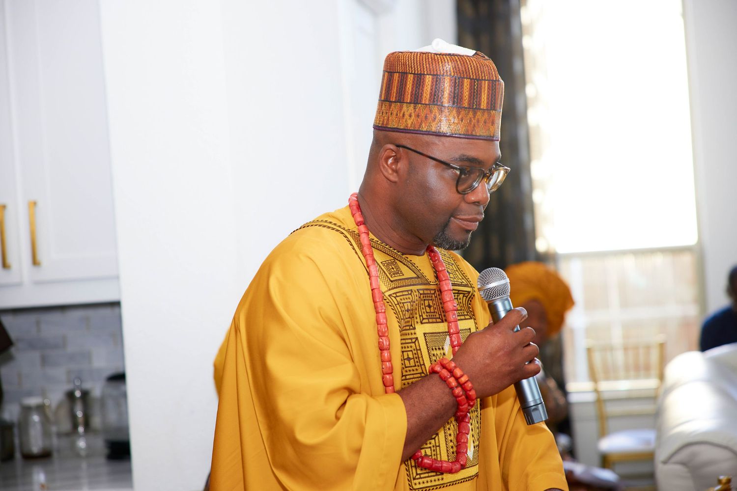 Person in traditional yellow African attire speaks into microphone during ceremonial event.