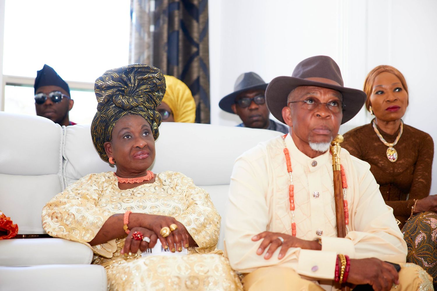 A traditional African gathering shows people seated together wearing bright ceremonial clothing and hats.