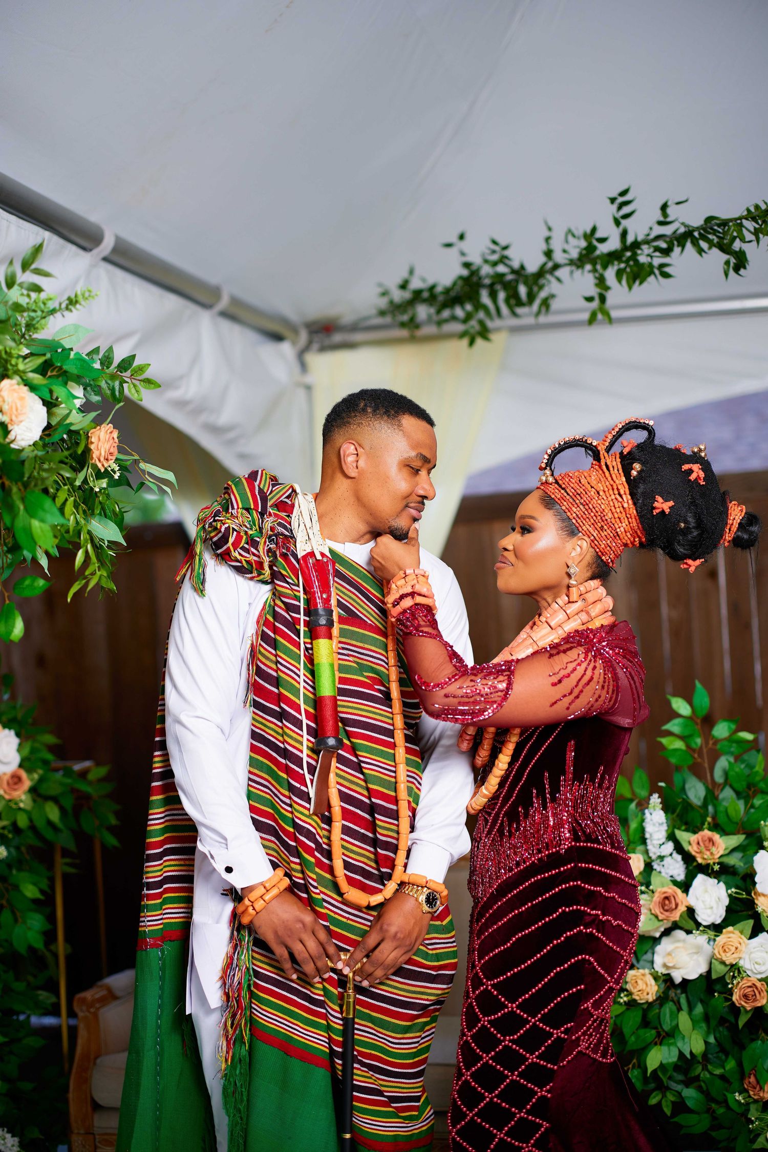 A couple in traditional African wedding attire embraces under a floral-decorated tent during a celebration.