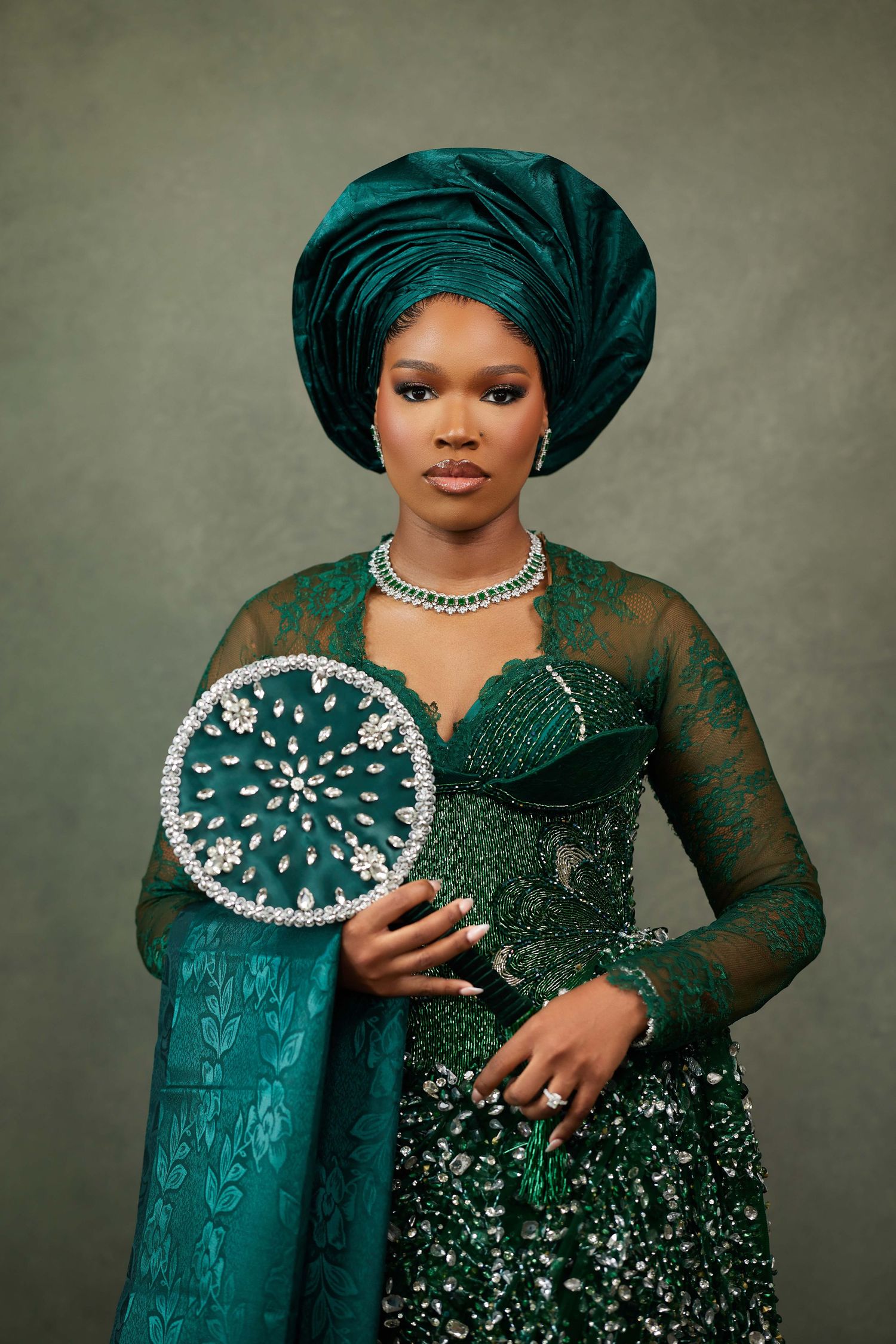 Portrait in emerald green lace dress with pearl accessories and matching headwrap against grey background.