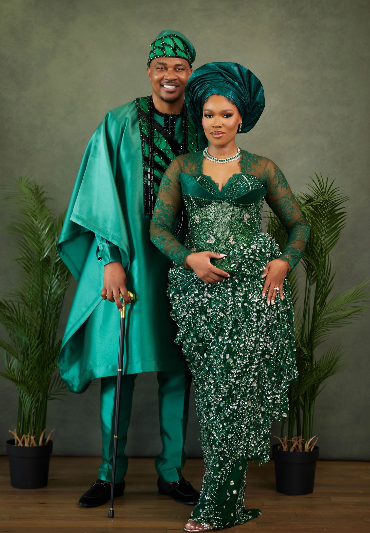 A couple in matching emerald green traditional Nigerian attire poses together in a studio setting with potted plants.