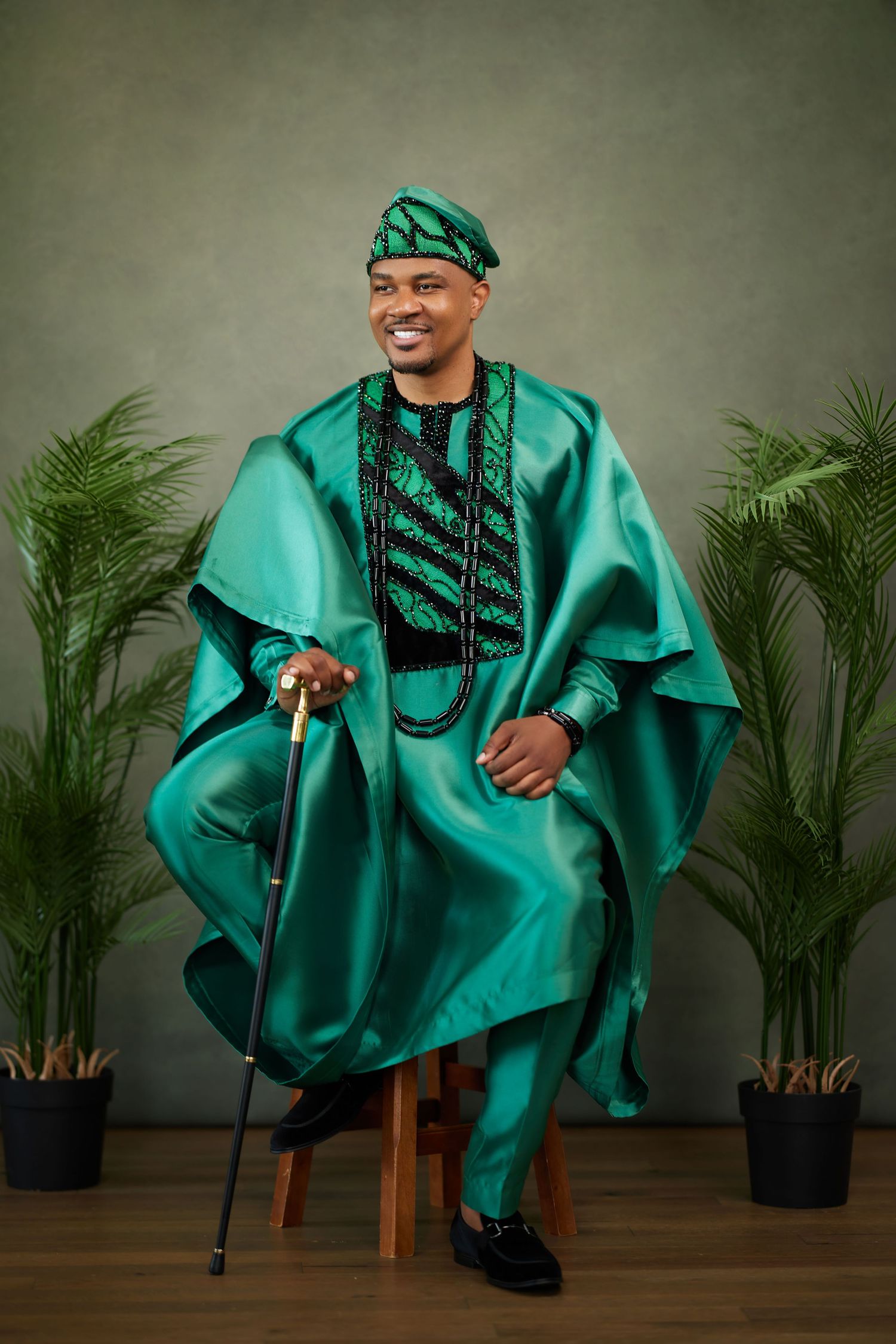 A person in a flowing emerald green agbada with black embellishments poses expressively against a dark backdrop with palm plants.