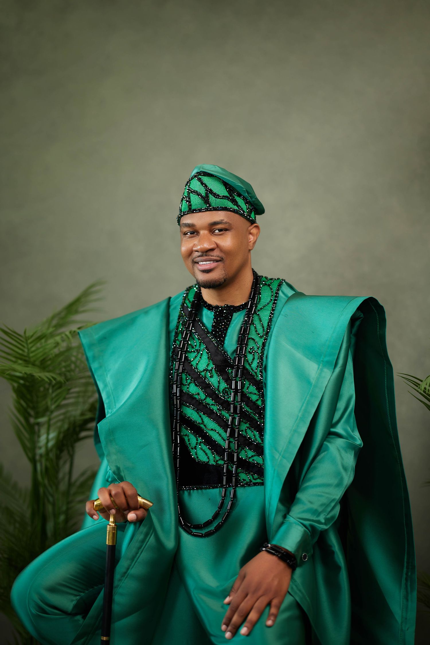 Traditional Nigerian formal attire in emerald green agbada with matching cap and black beaded necklaces against grey backdrop.
