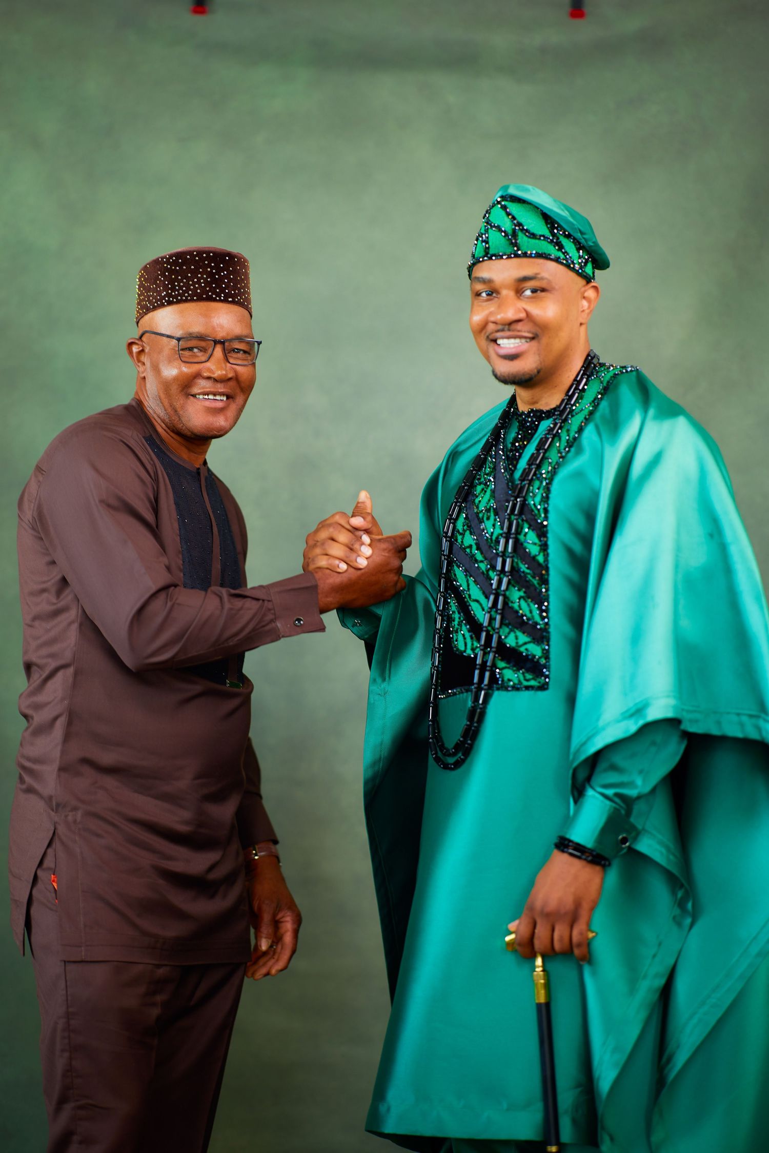 Two men in contrasting black and turquoise traditional attire share a joyful handshake during a formal gathering.