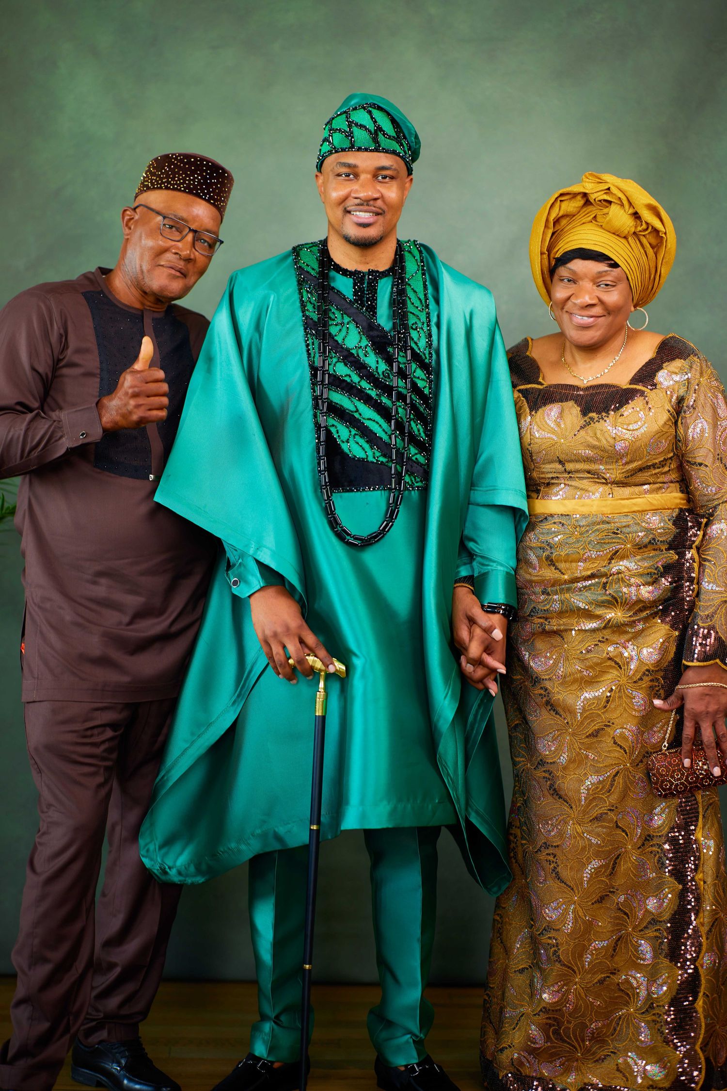 Three people in traditional Nigerian attire pose together, wearing green agbada, black outfit, and golden dress.