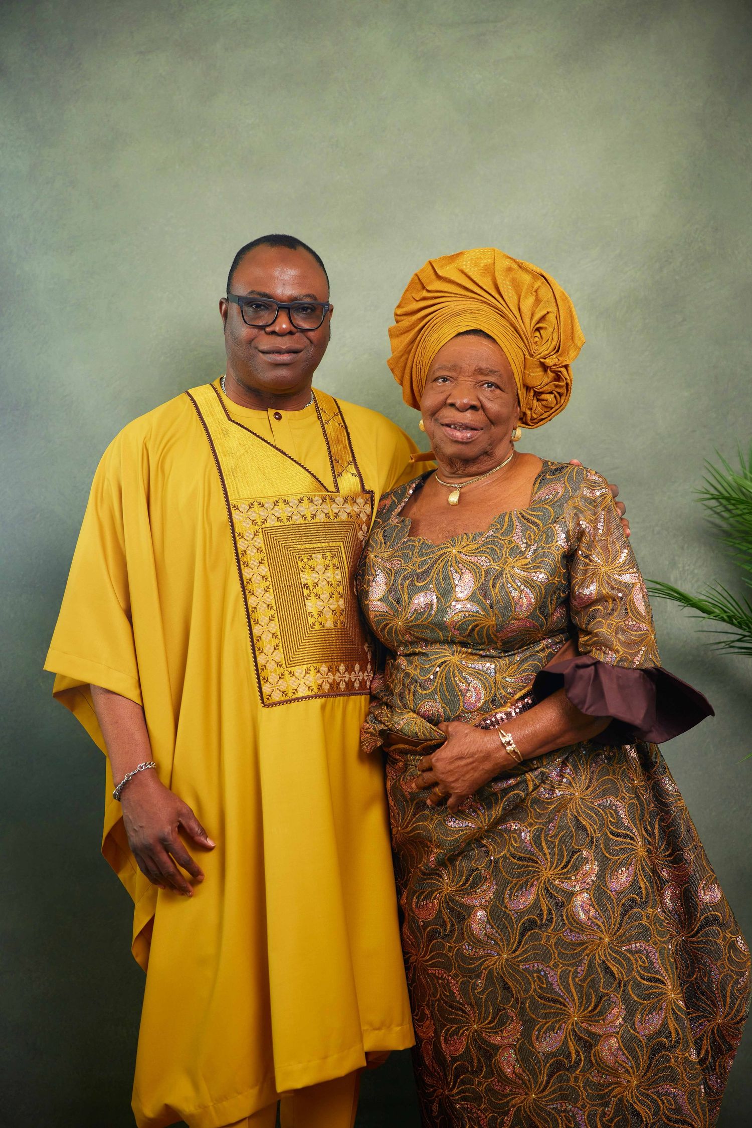Two people in traditional African attire pose together, wearing yellow and brown patterned clothing against a gray background.