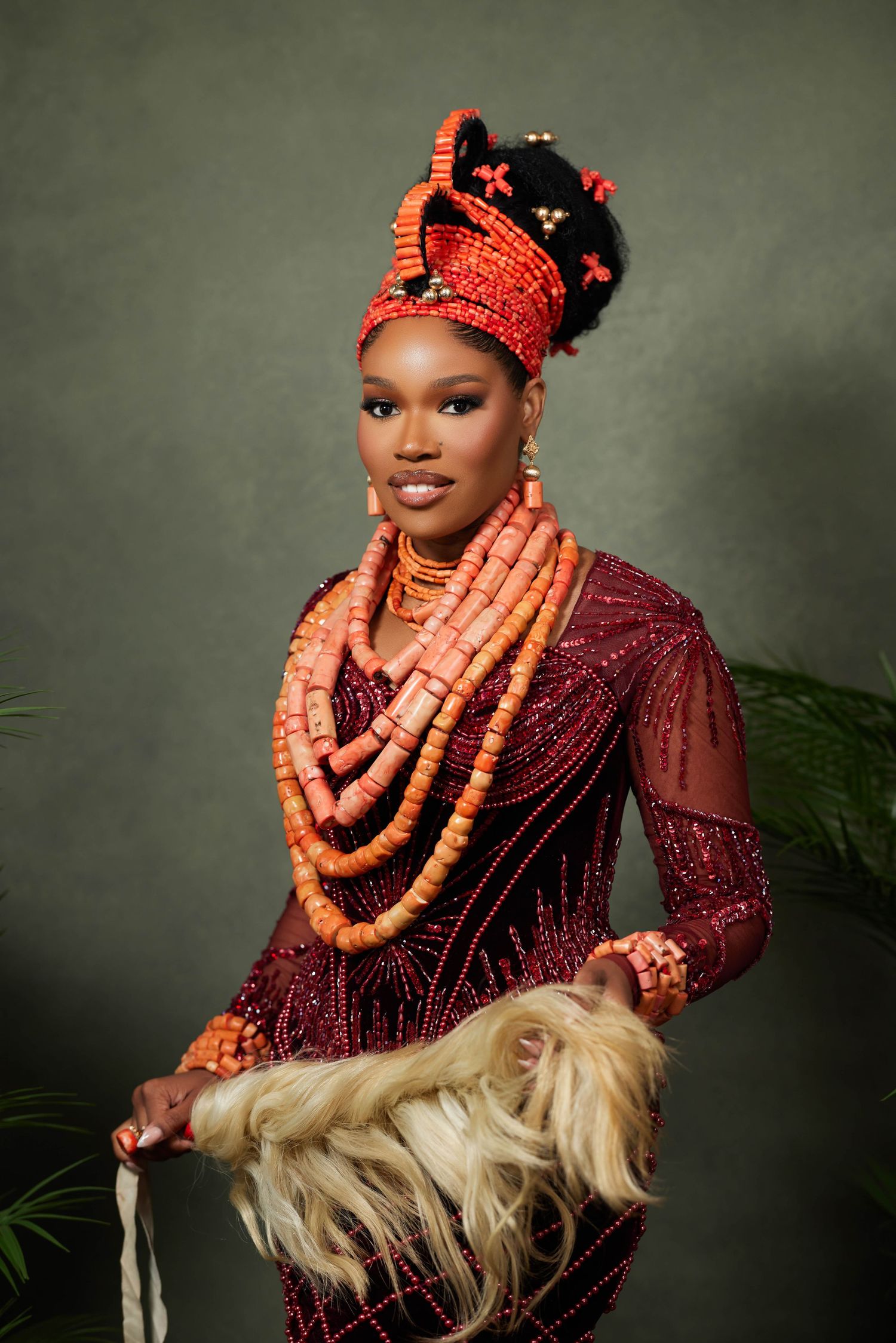 Traditional Nigerian portrait showing ornate coral beads, burgundy dress and decorative headdress against gray backdrop.