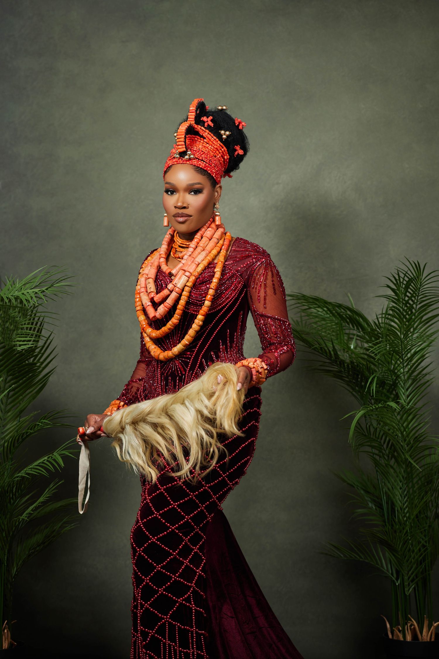 Traditional Nigerian bride wearing burgundy dress and coral beads poses elegantly against a moody background with palm plants.