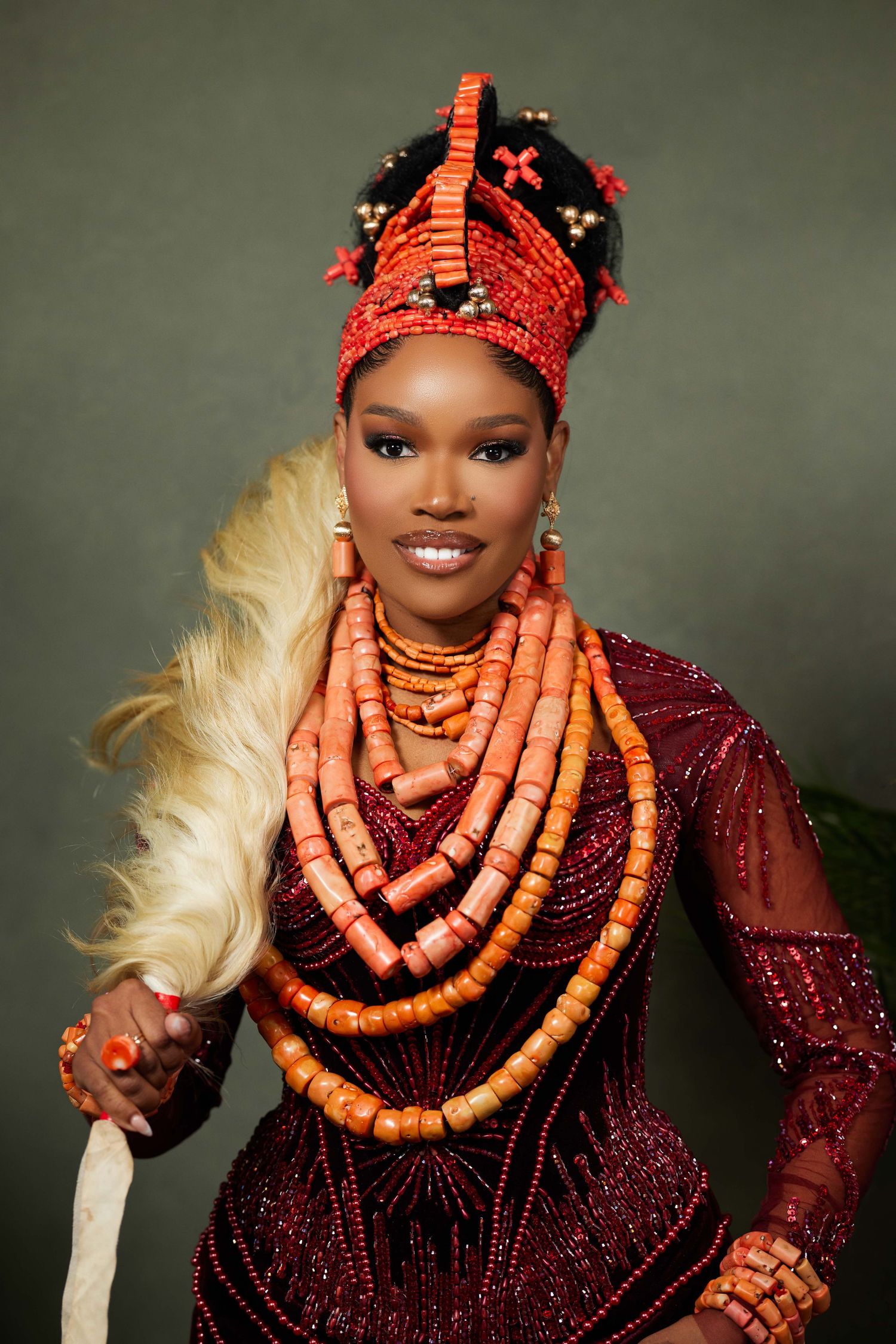 Traditional Nigerian wedding attire showcasing coral beaded necklaces and burgundy dress against dark background.