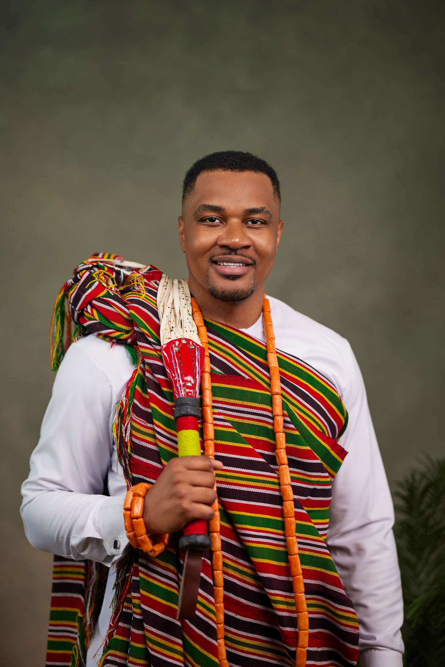 Individual in white shirt and colorful striped kente cloth accessory smiling in studio portrait.