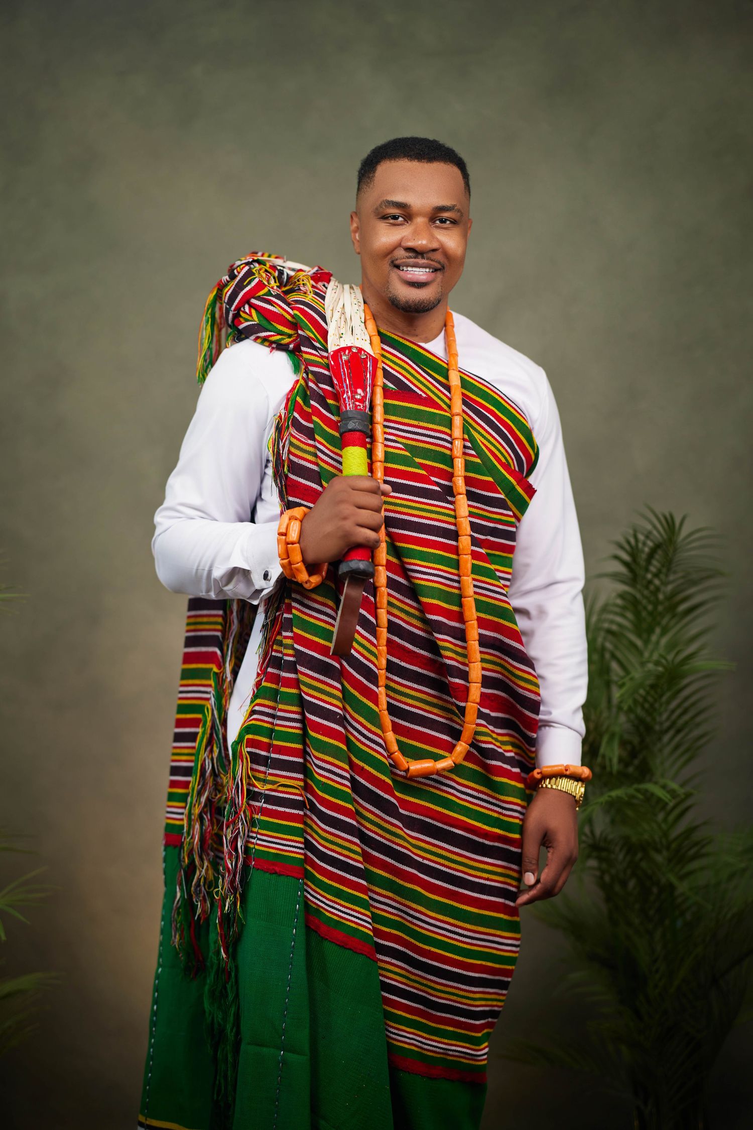 A person poses in traditional African attire featuring a colorful striped sash over a white shirt and green pants.