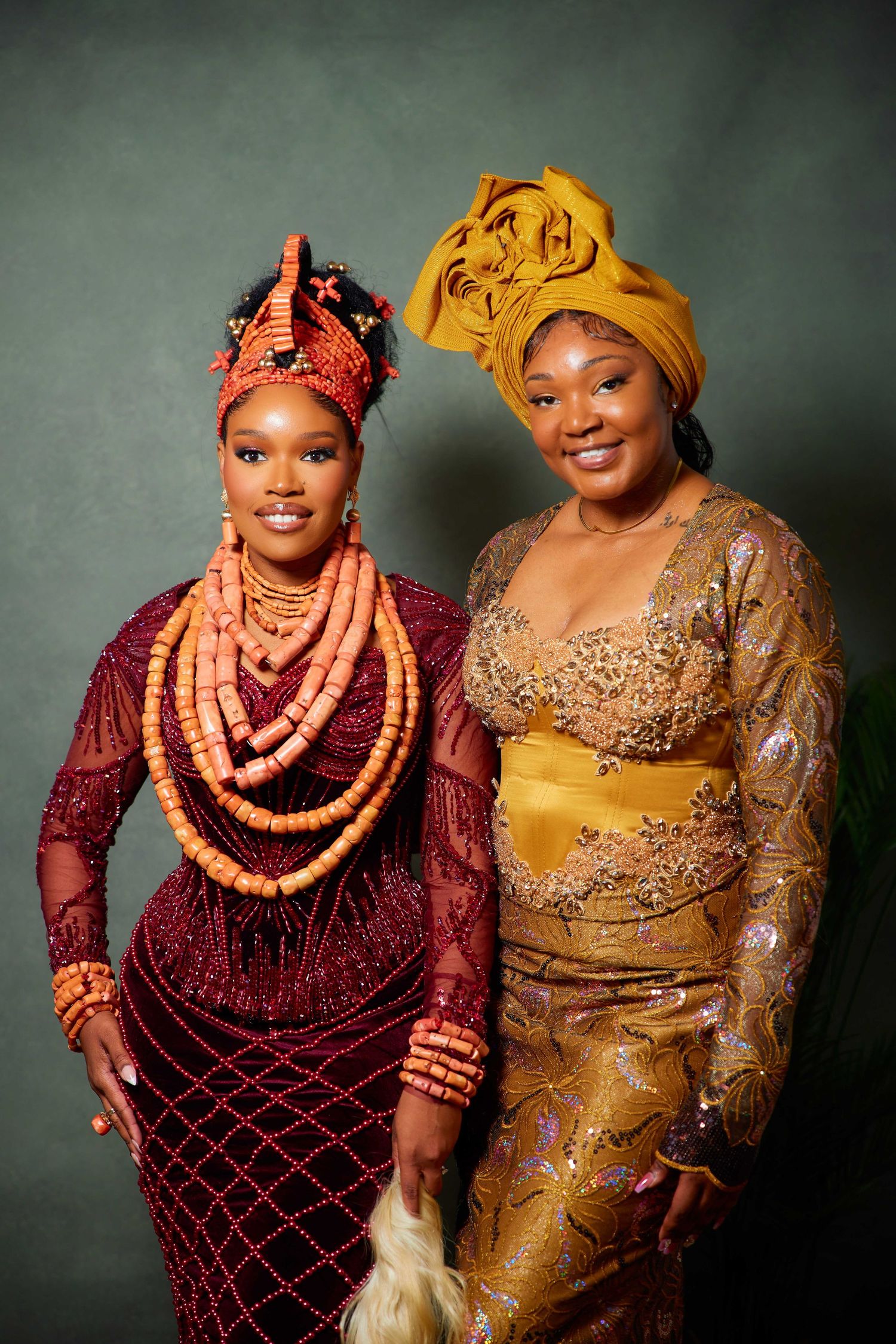 Two people pose in traditional Nigerian wedding attire featuring burgundy and gold colors with coral beaded jewelry.