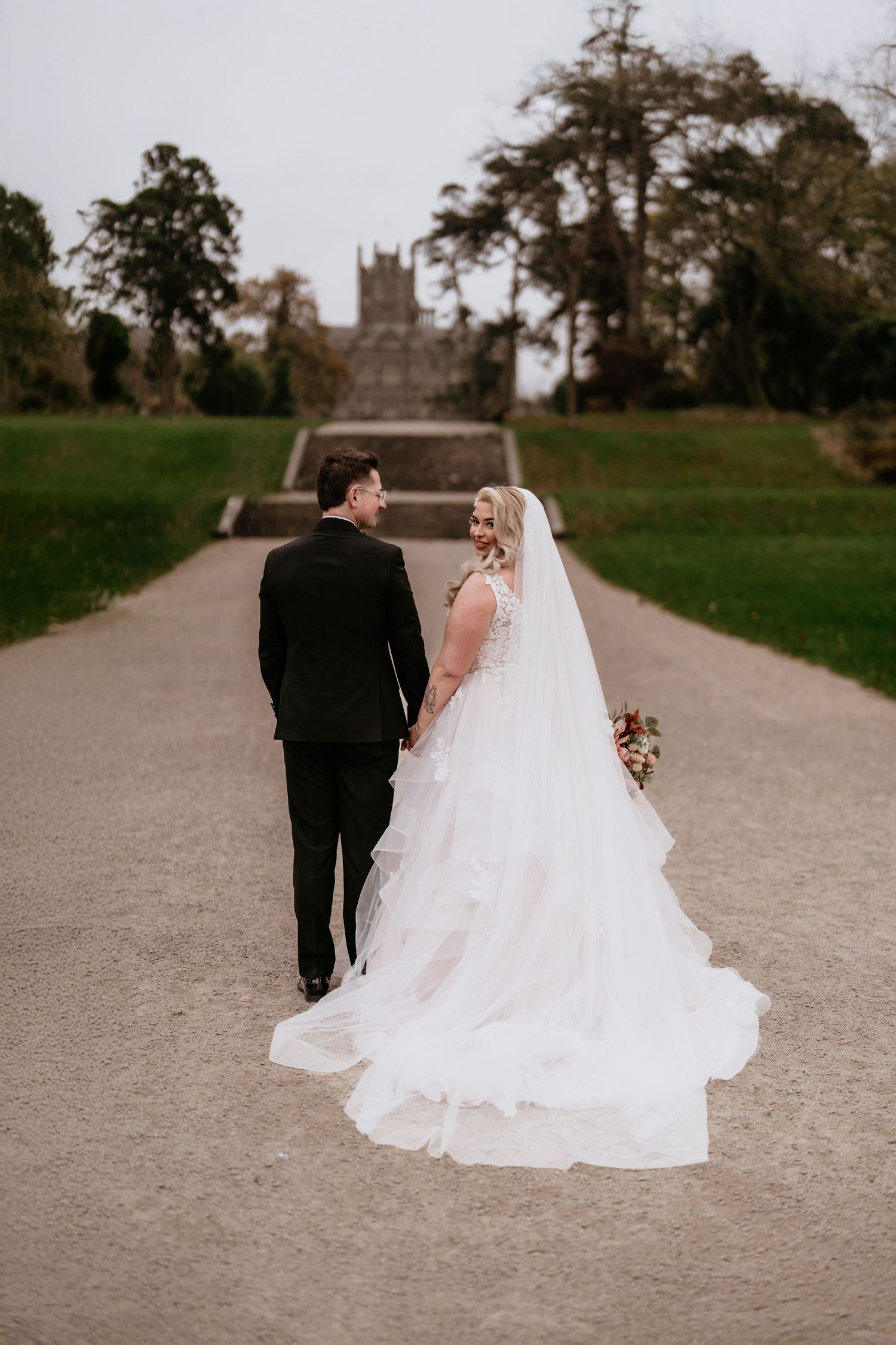 A couple in wedding attire walking along a path towards a historic castle building in The Orangery Margam Park