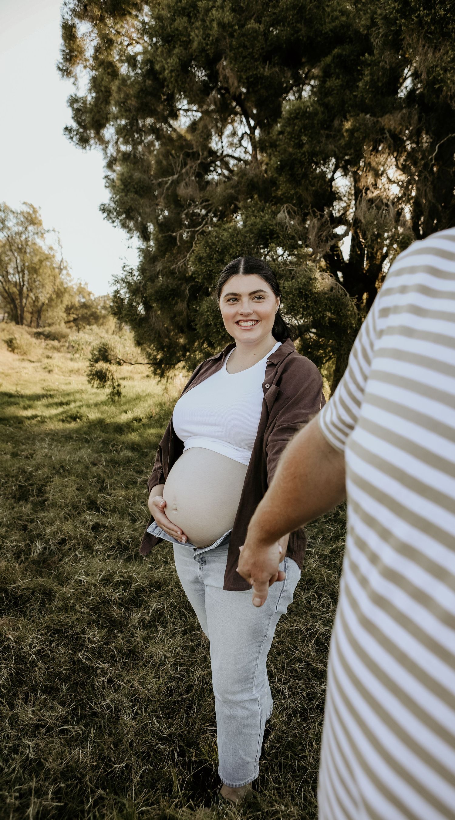 A pregnant person in a brown hoodie and white shirt stands outdoors in a natural setting during golden hour.