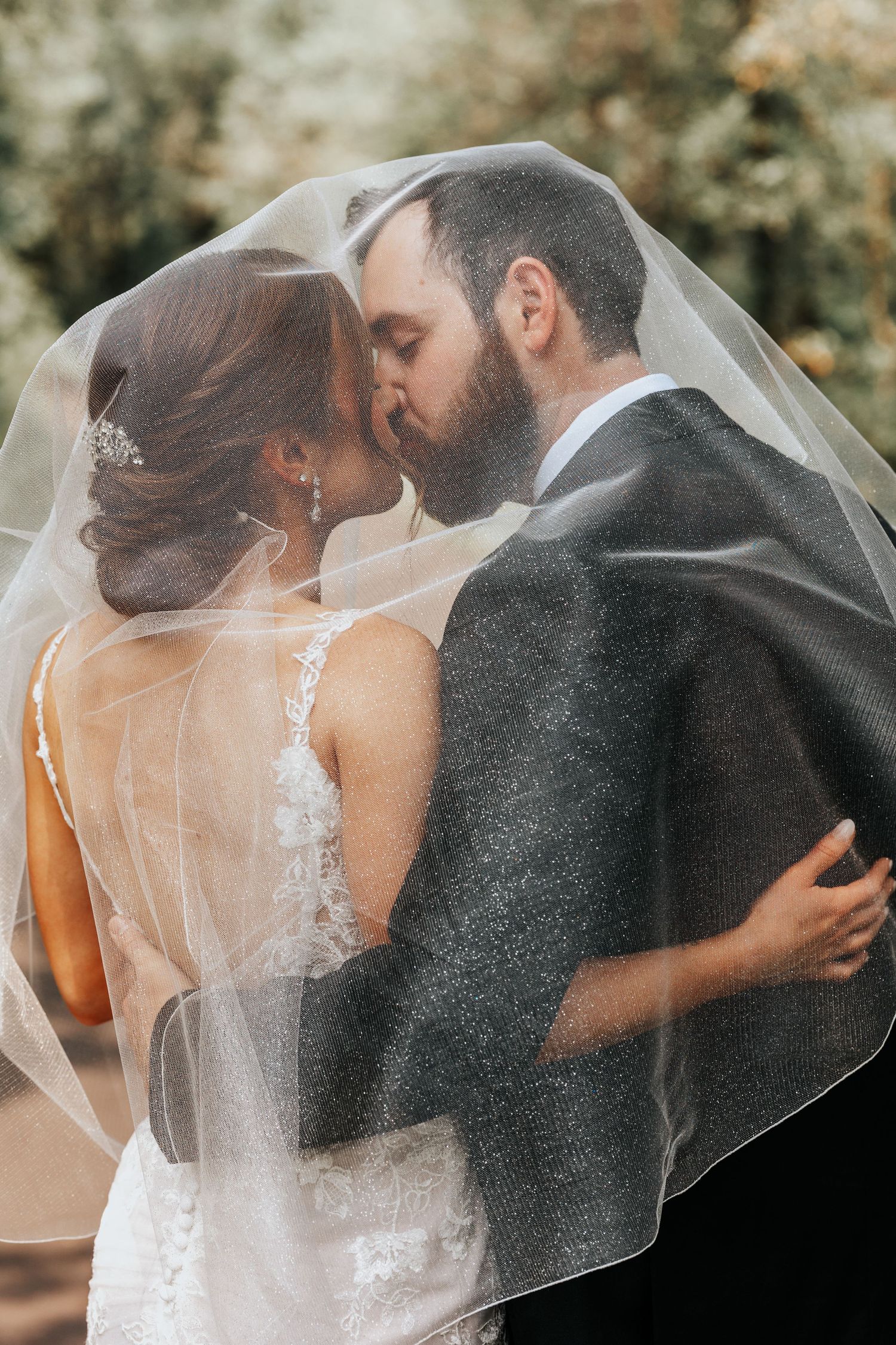 Romantic wedding couple shares intimate kiss under flowing white veil in outdoor garden setting.